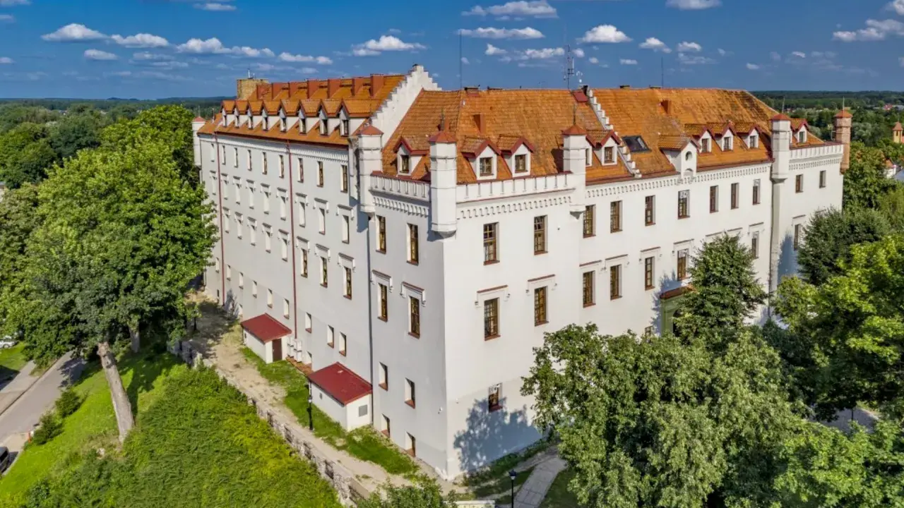 Biały zamek z czerwonym dachem, otoczony zielenią, oferuje wyjątkowe noclegi w zamkach i pałacach. Idealne miejsce na romantyczny wypad.