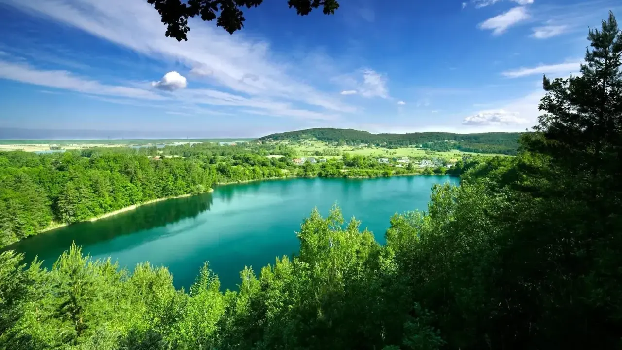 Lazurowe jezioro w Polsce, otoczone zielonymi lasami i wzgórzami, pod błękitnym niebem z białymi chmurami.