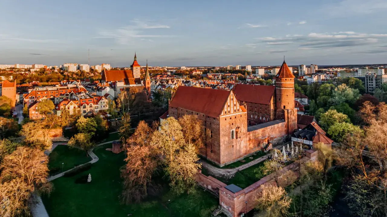 Zachód słońca nad zamkami krzyżackimi na Mazurach. Potężne ceglane mury i wieże w blasku złotej godziny, otoczone zielenią i miastem.