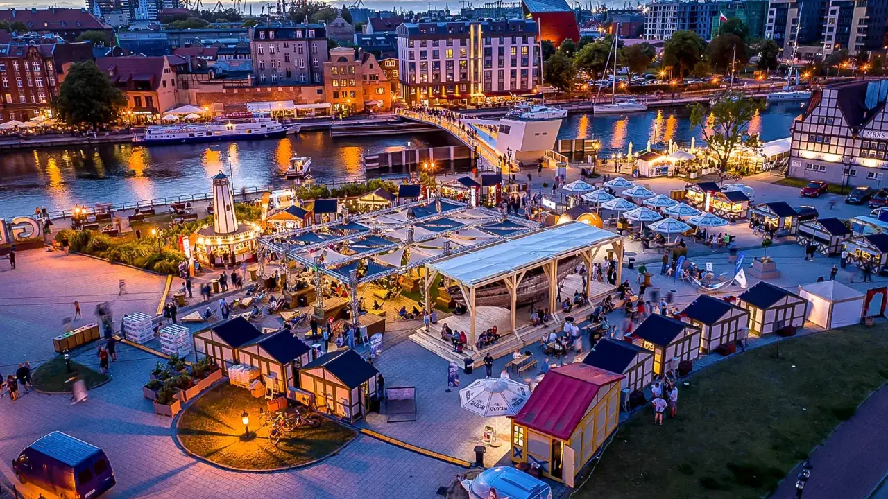 Tętniący życiem jarmark Gdańsk wieczorem. Widać rzekę Motławę, łodzie, most, liczne stoiska i ludzi spacerujących wśród iluminacji.