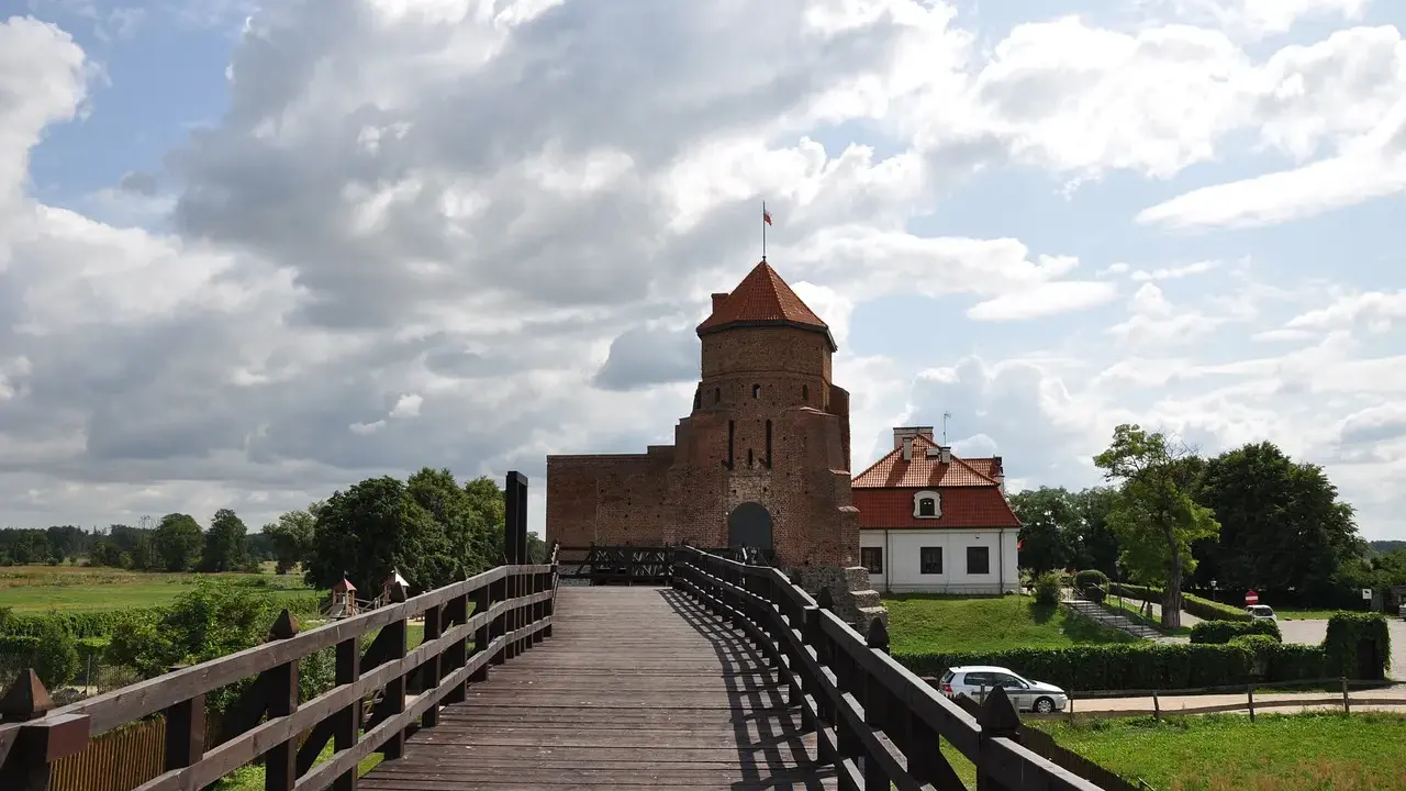 Drewniany most prowadzi do ceglanego zamku, jednego z najpiękniejszych zamków na Mazowszu. W tle biały budynek i zielone drzewa.