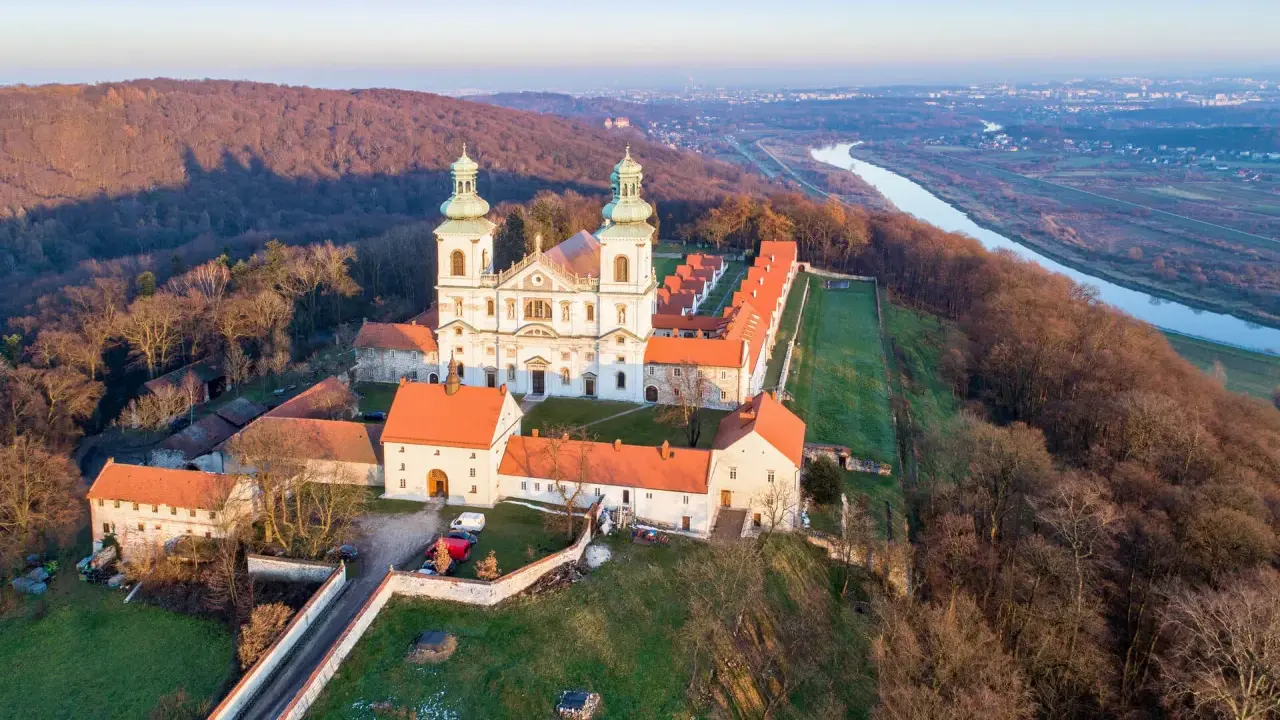 Wielki klasztor na wzgórzu, niczym milczący zakonnik, wita pierwsze promienie słońca. Obok rzeka i miasto w oddali.