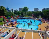 Tętniące życiem baseny odkryte pełne ludzi kąpiących się i bawiących w słońcu. W tle miejska panorama.