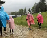 Rodzina w kolorowych pelerynach idzie ścieżką w **Zakopanem w deszczu**. W tle widać górski krajobraz i budynki.