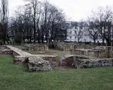 Ruiny zamku w Wieluniu, kamienne i ceglane mury na tle drzew i białego budynku.