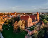 Zachód słońca nad zamkami krzyżackimi na Mazurach. Potężne ceglane mury i wieże w blasku złotej godziny, otoczone zielenią i miastem.