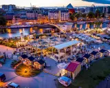 Tętniący życiem jarmark Gdańsk wieczorem. Widać rzekę Motławę, łodzie, most, liczne stoiska i ludzi spacerujących wśród iluminacji.