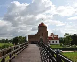 Drewniany most prowadzi do ceglanego zamku, jednego z najpiękniejszych zamków na Mazowszu. W tle biały budynek i zielone drzewa.