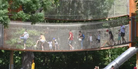 Wesoły park linowy dla dzieci z mostkami i zjeżdżalnią. Dorośli i dzieci bawią się na wysokości, ciesząc się letnim dniem.