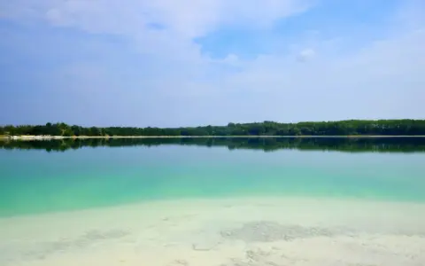 Lazurowe jezioro z piaszczystym dnem kusi do kąpieli. Piękny, spokojny krajobraz z zielonym lasem w tle.