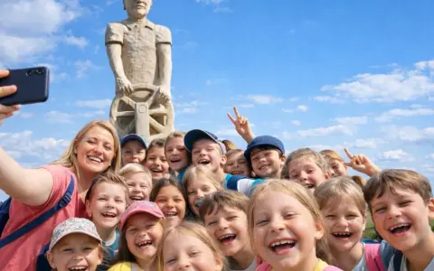 Uśmiechnięta grupa dzieci i opiekunka robią sobie selfie pod drewnianym pomnikiem. Wspaniałe kościerzyna atrakcje dla dzieci!