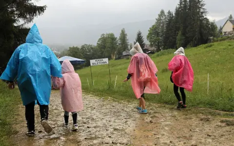 Rodzina w kolorowych pelerynach idzie ścieżką w **Zakopanem w deszczu**. W tle widać górski krajobraz i budynki.