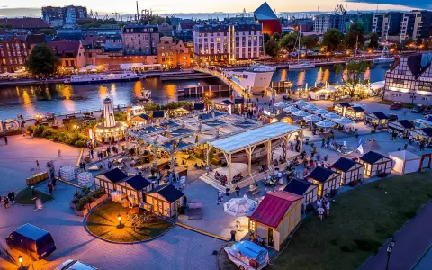 Tętniący życiem jarmark Gdańsk wieczorem. Widać rzekę Motławę, łodzie, most, liczne stoiska i ludzi spacerujących wśród iluminacji.