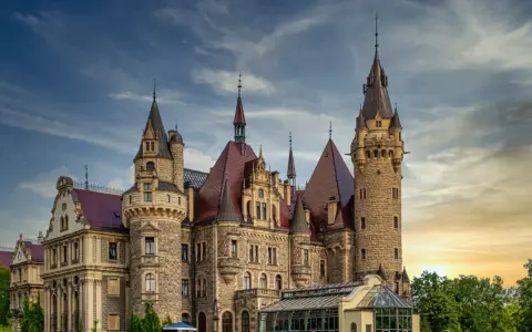 Imponujący zamek w Polsce z kamienia, z wieloma wieżami i czerwonymi dachami, na tle dramatycznego nieba o zachodzie słońca.