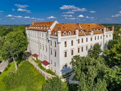 Biały zamek z czerwonym dachem, otoczony zielenią, oferuje wyjątkowe noclegi w zamkach i pałacach. Idealne miejsce na romantyczny wypad.