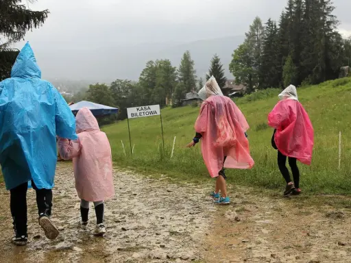 Rodzina w kolorowych pelerynach idzie ścieżką w **Zakopanem w deszczu**. W tle widać górski krajobraz i budynki.