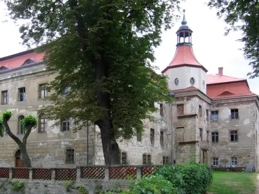 Zamek w Domanicach, zniszczony, z czerwoną dachem i wieżą, otoczony zielenią.