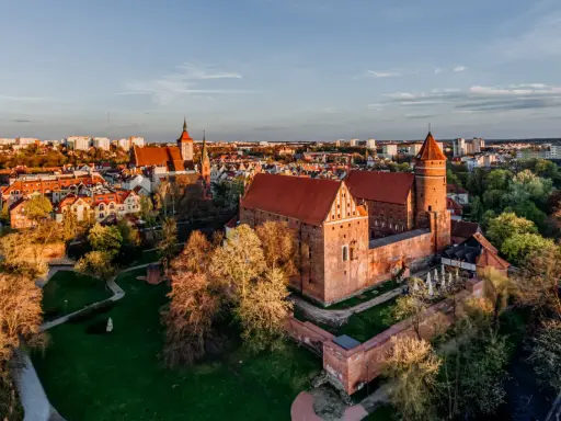 Zachód słońca nad zamkami krzyżackimi na Mazurach. Potężne ceglane mury i wieże w blasku złotej godziny, otoczone zielenią i miastem.