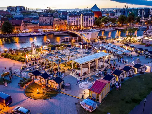 Tętniący życiem jarmark Gdańsk wieczorem. Widać rzekę Motławę, łodzie, most, liczne stoiska i ludzi spacerujących wśród iluminacji.