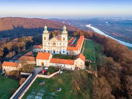 Wielki klasztor na wzgórzu, niczym milczący zakonnik, wita pierwsze promienie słońca. Obok rzeka i miasto w oddali.