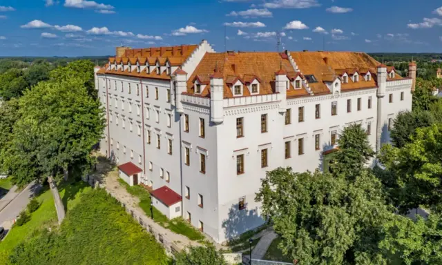 Biały zamek z czerwonym dachem, otoczony zielenią, oferuje wyjątkowe noclegi w zamkach i pałacach. Idealne miejsce na romantyczny wypad.