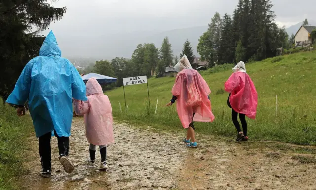 Rodzina w kolorowych pelerynach idzie ścieżką w **Zakopanem w deszczu**. W tle widać górski krajobraz i budynki.