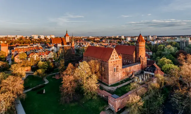 Zachód słońca nad zamkami krzyżackimi na Mazurach. Potężne ceglane mury i wieże w blasku złotej godziny, otoczone zielenią i miastem.