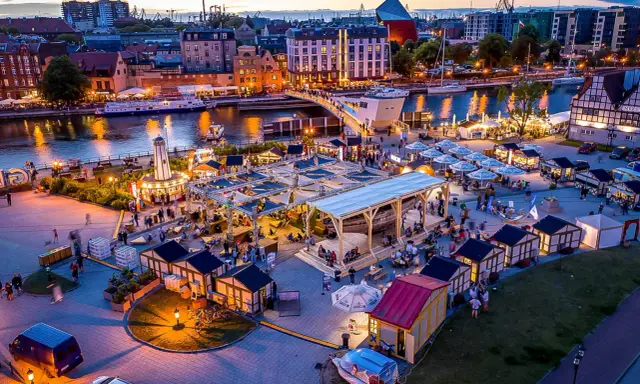 Tętniący życiem jarmark Gdańsk wieczorem. Widać rzekę Motławę, łodzie, most, liczne stoiska i ludzi spacerujących wśród iluminacji.