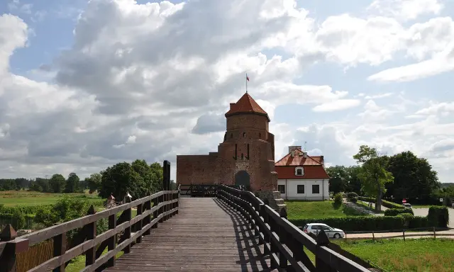 Drewniany most prowadzi do ceglanego zamku, jednego z najpiękniejszych zamków na Mazowszu. W tle biały budynek i zielone drzewa.