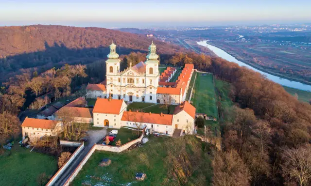 Wielki klasztor na wzgórzu, niczym milczący zakonnik, wita pierwsze promienie słońca. Obok rzeka i miasto w oddali.