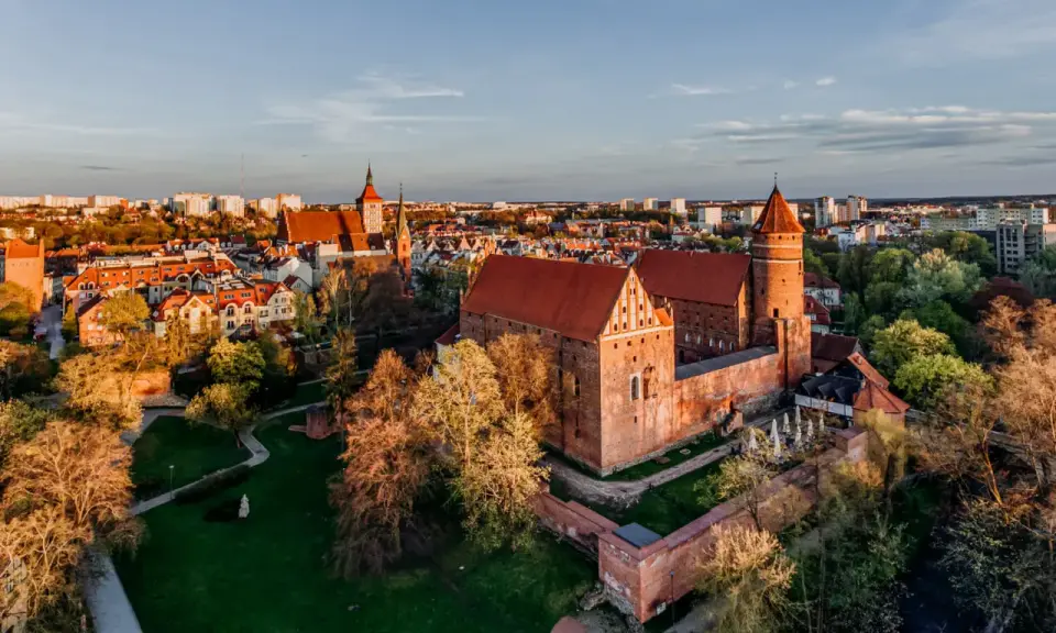 Zachód słońca nad zamkami krzyżackimi na Mazurach. Potężne ceglane mury i wieże w blasku złotej godziny, otoczone zielenią i miastem.