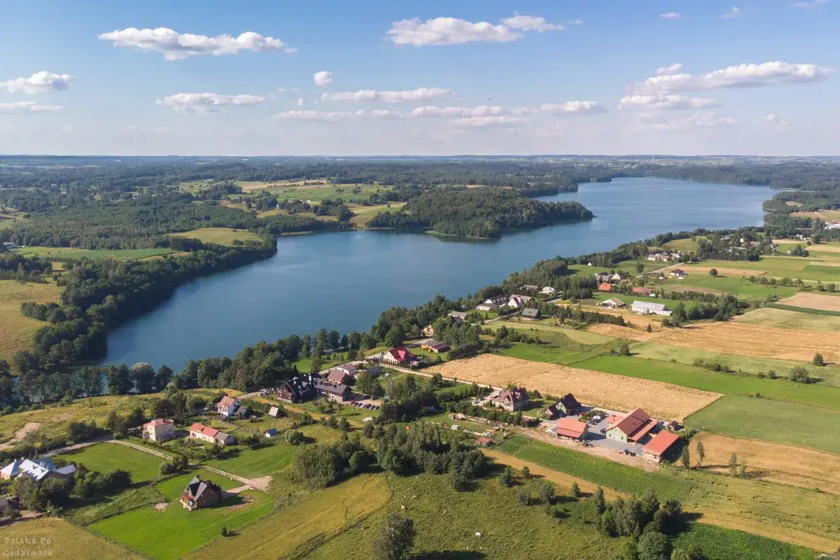 Malowniczy krajobraz z jeziorem najgłębszym w Polsce, otoczonym lasami i polami. W oddali widać zabudowania.
