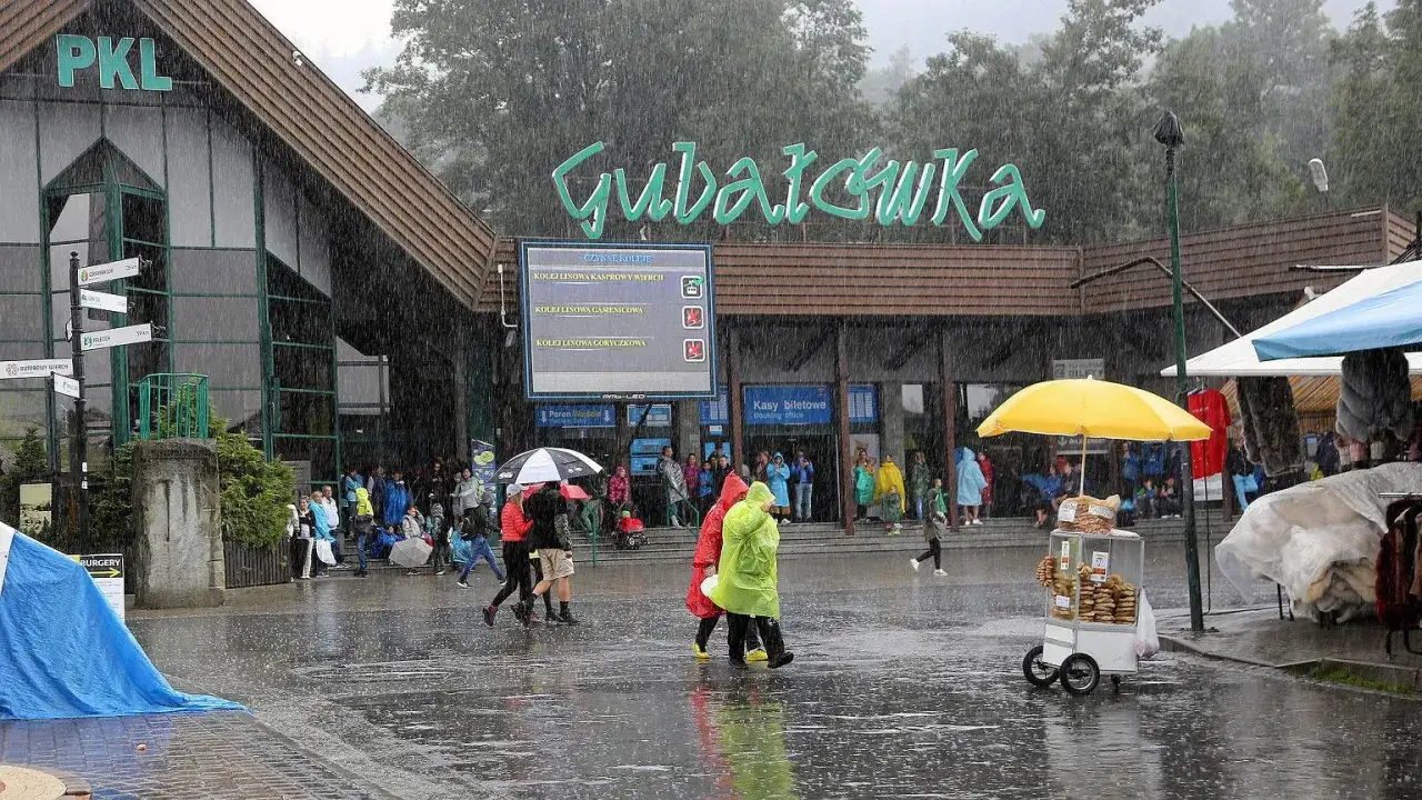 Ludzie w kolorowych płaszczach przeciwdeszczowych i pod parasolami spieszą się pod Gubałówkę w deszczu. Mokry chodnik odbija światła.