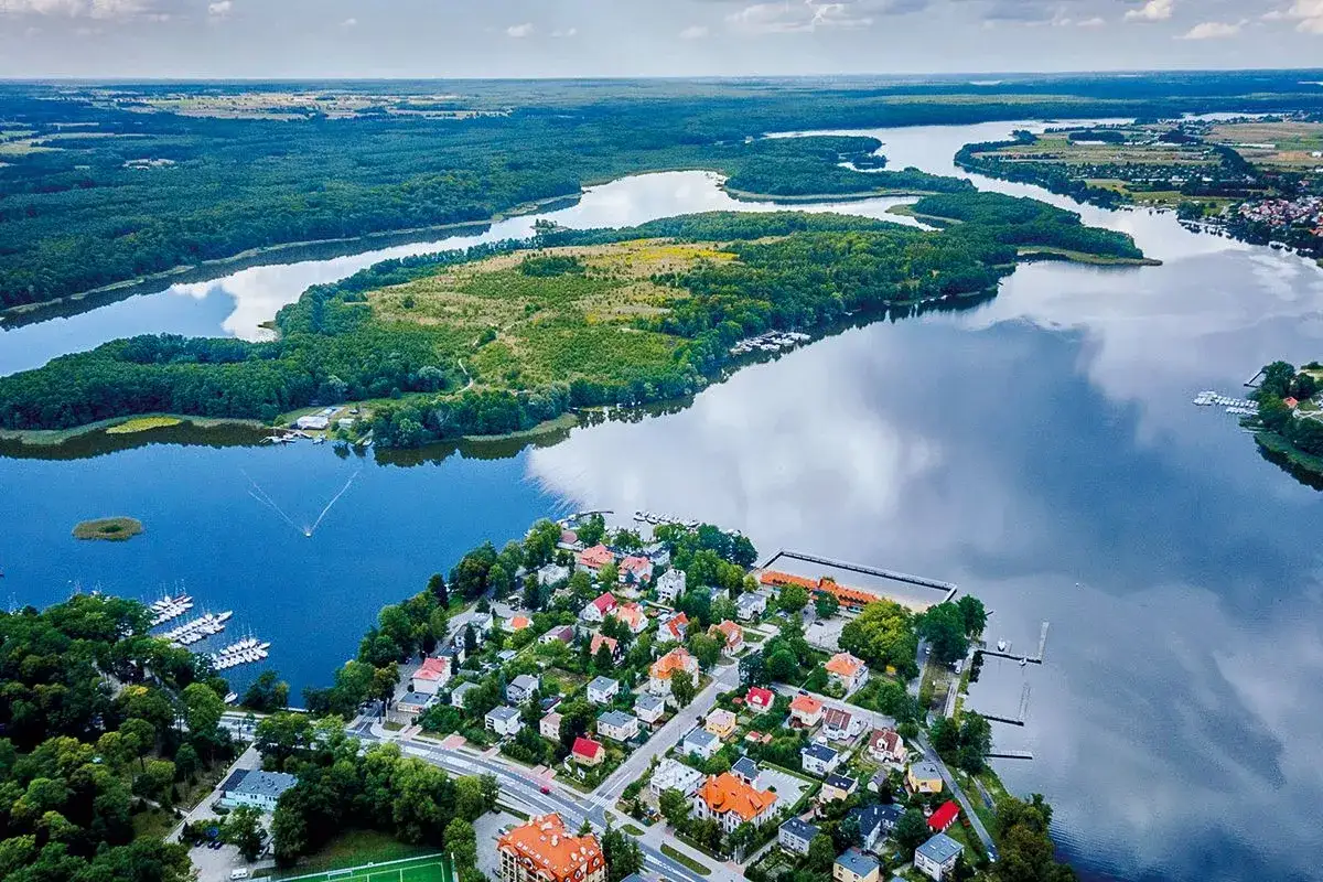 Malowniczy widok na krainę tysiąca jezior. Widać miasteczko z kolorowymi domami, przystań z łodziami i rozległe, leśne tereny nad wodą.