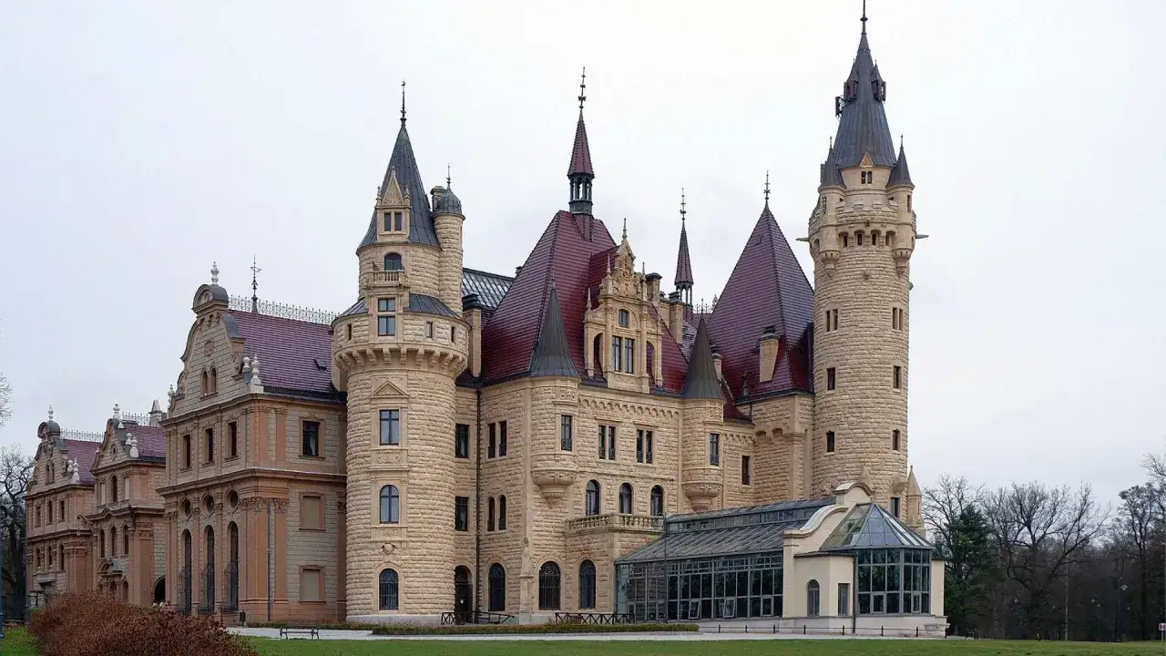 Kamienny pałac z czerwonymi dachami i wieżami, największy pałac w Polsce, otoczony zielenią.