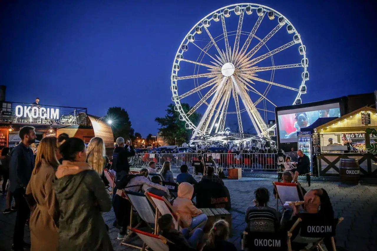 Wiecz&oacute;r na jarmarku dominikańskim: ludzie oglądają film podświetlanym kołem widokowym i stoiskami z jedzeniem.
