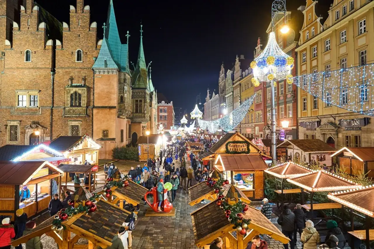 Tłum ludzi na największym jarmarku bożonarodzeniowym w Polsce, wśród drewnianych straganów i świątecznych iluminacji.
