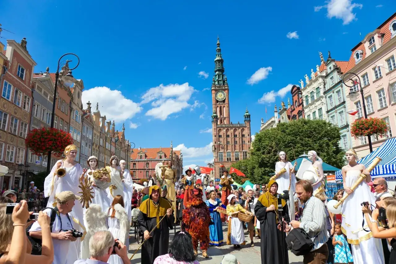 Kolorowy jarmark Gdańsk z postaciami na szczudłach i tłumem widzów na tle zabytkowych kamienic.