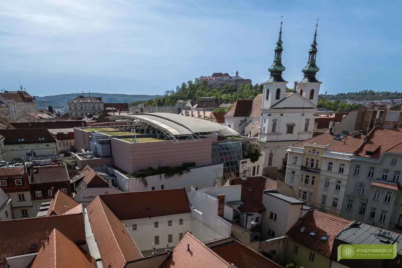 Widok na dachy Brna, z kościołem o dwóch wieżach i zamkiem na wzgórzu. Wśród miejskich atrakcji wyróżnia się nowoczesny budynek z zielonym dachem.