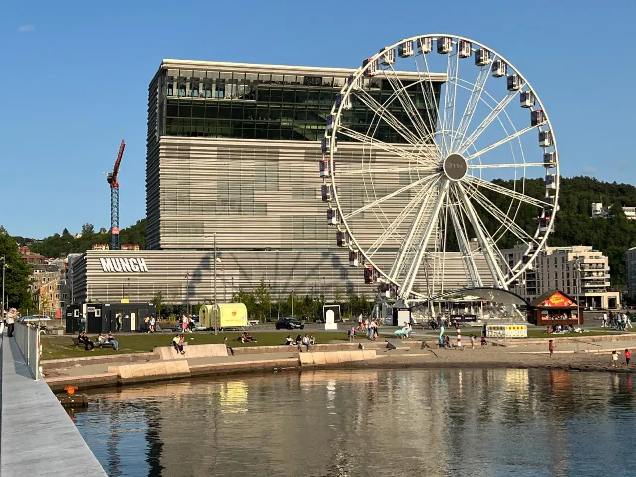 Oslo atrakcje: Muzeum Muncha i diabelski młyn na nabrzeżu, gdzie ludzie odpoczywają nad wodą.