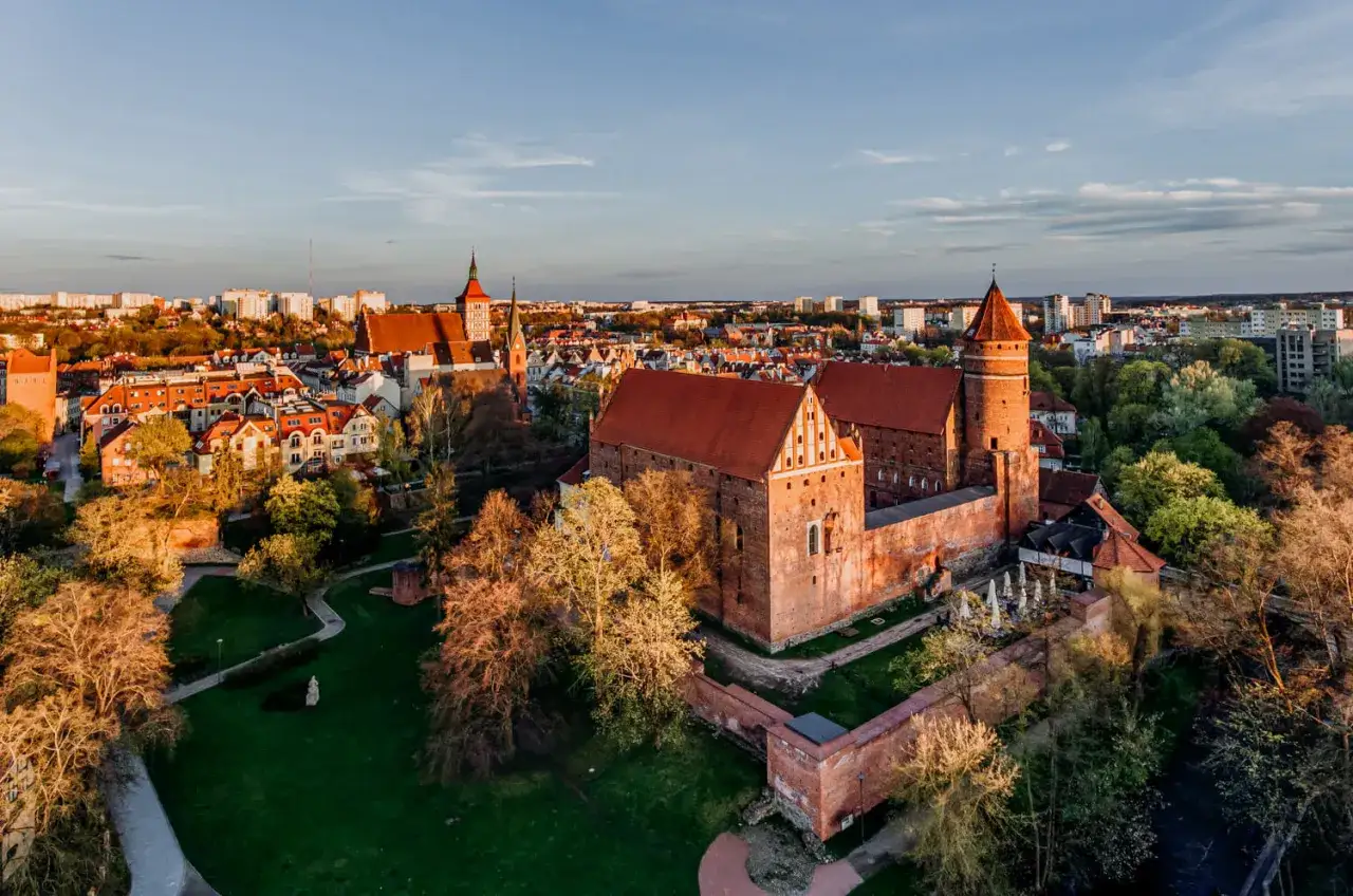 Zach&oacute;d słońca nad zamkami krzyżackimi na Mazurach. Potężne mury i wieże w ciepłym świetle, otoczone zielenią i miastem.