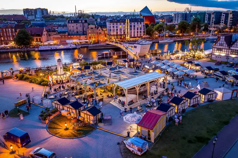 Tętniący życiem jarmark Gdańsk wieczorem. Widać rzekę Motławę, łodzie, most, liczne stoiska i ludzi spacerujących wśród iluminacji.